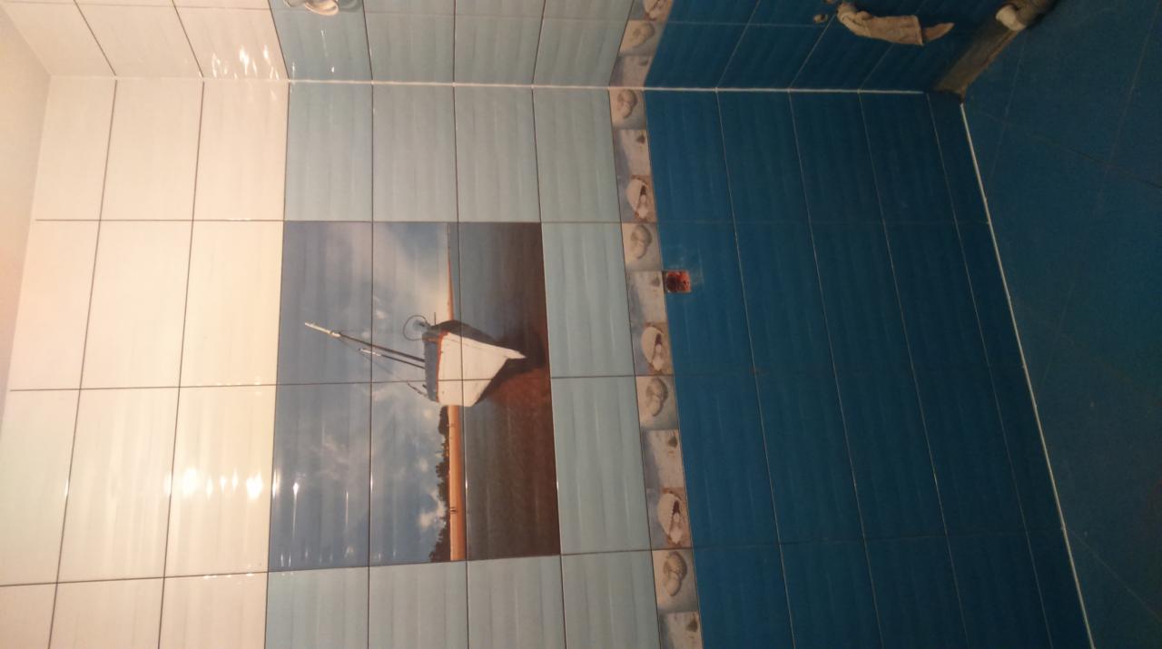 Wykończona ściana łazienki z białymi, jasnoniebieskimi i ciemnoniebieskimi płytkami. Na jasnoniebieskich płytkach dekoracyjny motyw żaglówki na plaży.