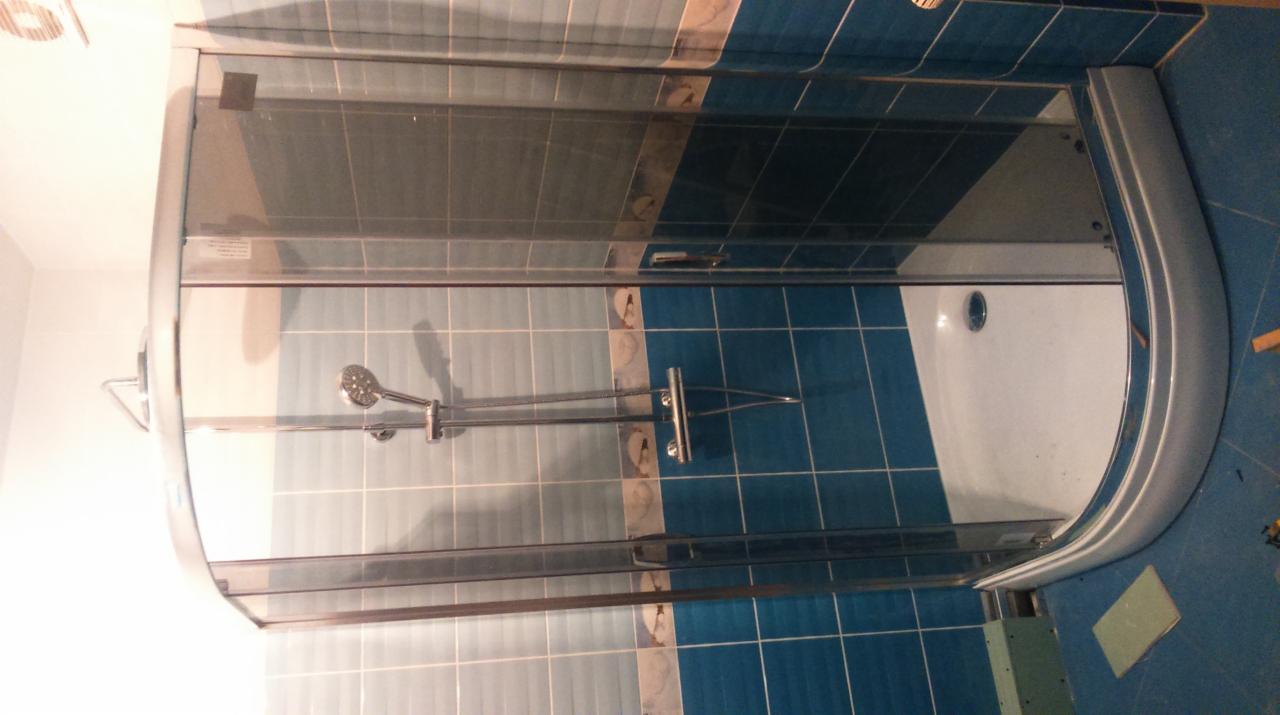 Zamknięta kabina prysznicowa z chromowanymi okuciami i szklanymi drzwiami, zamontowana w łazience z niebiesko-białymi płytkami, widok z góry z elementami niedokończonego montażu.