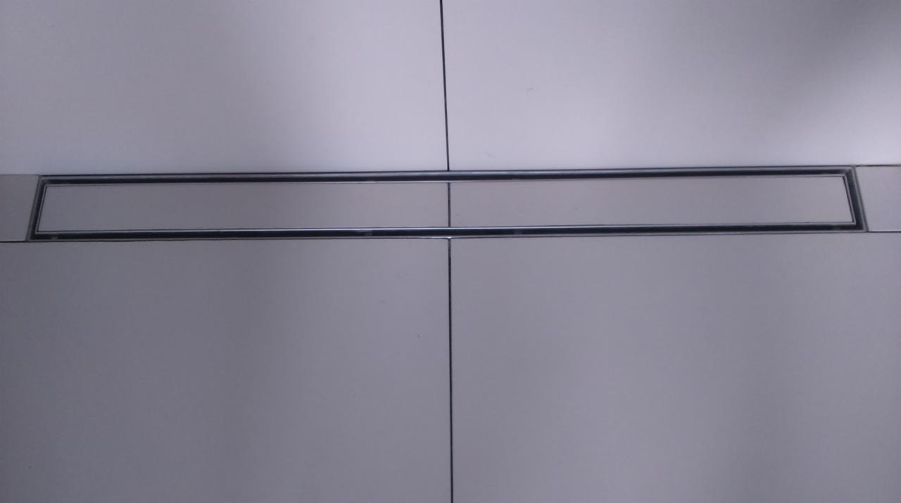 Szczegółowy widok liniowego odpływu prysznicowego z metalową ramką, wkomponowanego w jasne, kwadratowe płytki ceramiczne, z widocznymi fugami.