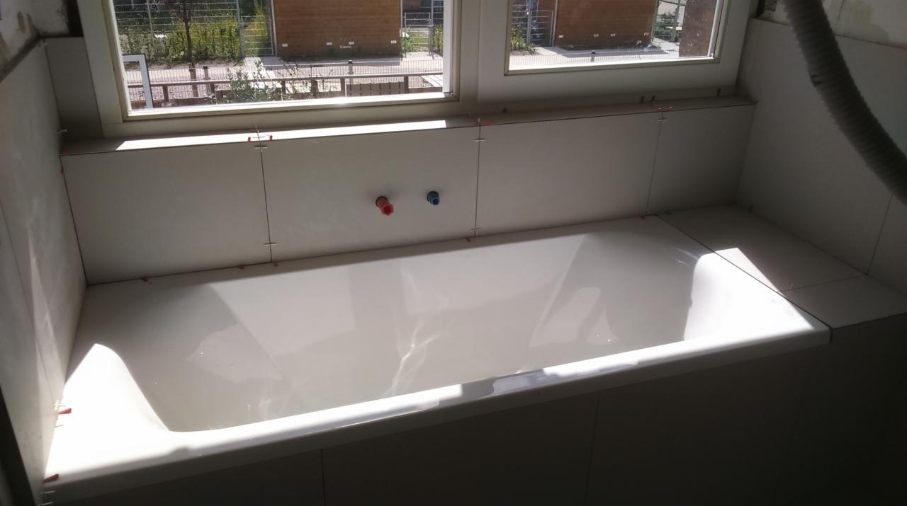 Biała wanna obudowana jasnymi płytkami w trakcie montażu, widoczne krzyżyki dystansowe, okno z widokiem na budynki w tle, przygotowane przyłącza wodne.
