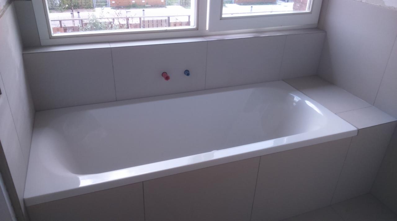 Biała wanna obudowana płytkami w trakcie montażu, widoczne przyłącza ciepłej i zimnej wody, okno w tle.