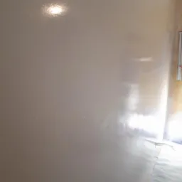 Świeżo nałożona, błyszcząca, beżowa warstwa na ścianie w trakcie remontu, z widocznymi kablami w dolnym rogu i odbiciem światła na powierzchni.