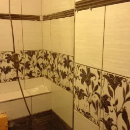 Fragment łazienki w trakcie wykańczania, widoczne płytki ścienne w dw&oacute;ch odcieniach beżu, z motywem kwiatowym i brązowym pasem, niezakończone fugowanie, wystający kabel elektryczny.