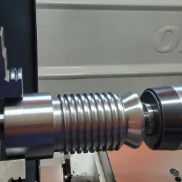 Metalowy wałek z wyżłobieniami obrabiany na tokarce, widoczne narzędzie skrawające i podziałka na elemencie mocującym.