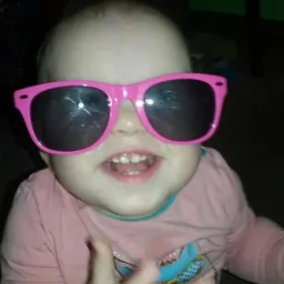 Uśmiechnięte dziecko w różowych okularach przeciwsłonecznych, z widocznymi pierwszymi ząbkami i wyciągniętą rączką, na rozmytym tle wnętrza.