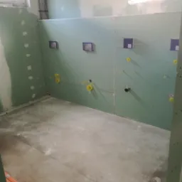 Wnętrze łazienki w trakcie montażu płyt kartonowo-gipsowych, widoczne otwory na instalacje sanitarne i elektryczne, surowy beton na podłodze.