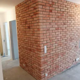 Wykończony narożnik ściany obłożony dekoracyjną cegłą w odcieniach czerwieni i brązu, z widocznymi gniazdkami elektrycznymi i włącznikiem światła, na tle surowego wnętrza w trakcie prac...