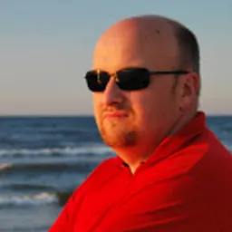 Mężczyzna w czerwonej koszulce i okularach przeciwsłonecznych na tle morza o zachodzie słońca. Portret z lekkim uśmiechem, ciepłe światło.