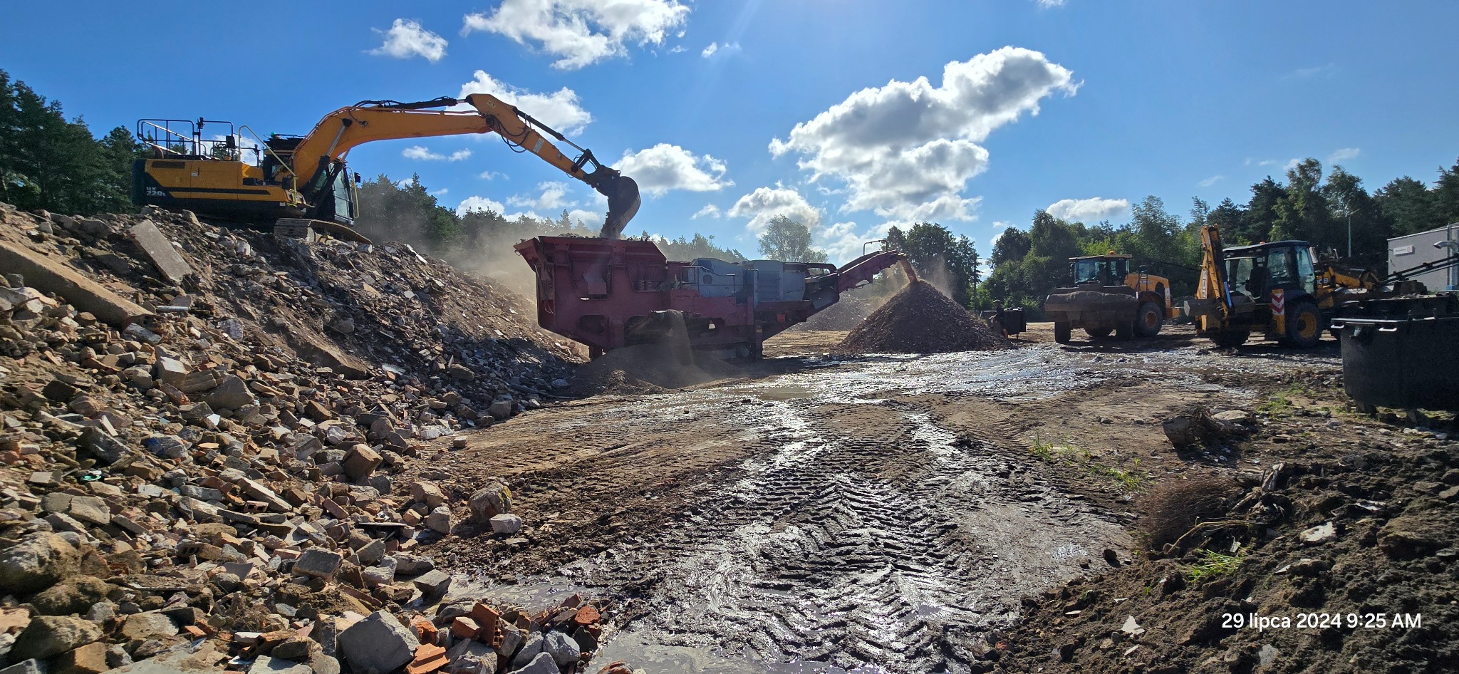 Żółta koparka podnosi gruz i wrzuca do czerwonego kruszarki, tworząc kopiec pokruszonego materiału na placu budowy z błotnistą ziemią i innymi maszynami w tle, pod błękitnym niebem z chmurami.
