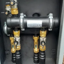 Rozdzielacz instalacji grzewczej w czarnej obudowie z zaworami, rurami i złączkami mosiężnymi, zamontowany przy ceglanej ścianie zewnętrznej, widoczne oznaczenia producenta.