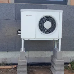 Biała jednostka zewnętrzna z logo Stiebel Eltron, zamontowana na metalowych wspornikach i betonowych bloczkach przy szarej ścianie budynku z oknem.