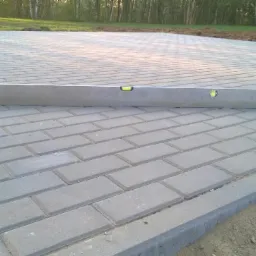 Układanie szarej kostki brukowej z użyciem poziomicy, widoczny fragment ułożonej nawierzchni i przygotowane podłoże.