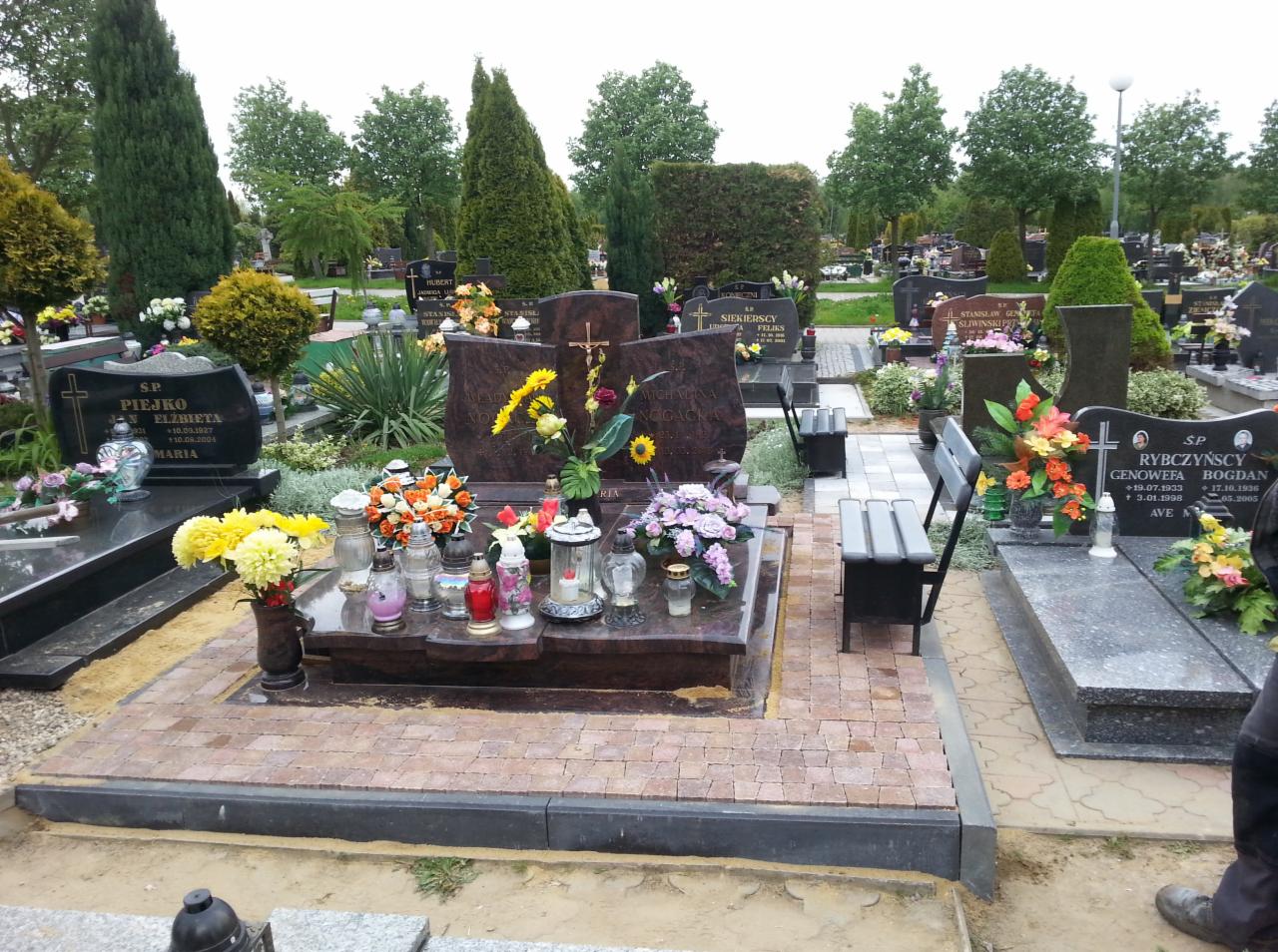 Świeżo ułożona kostka brukowa wokół nagrobka z kwiatami i zniczami na cmentarzu, widoczne inne groby i drzewa w tle.