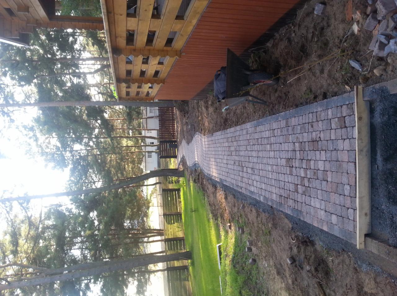 Kręta ścieżka z kostki brukowej w odcieniach brązu prowadząca wzdłuż drewnianego budynku, obrzeża wykończone drobnym żwirem, w tle widoczne drzewa i trawnik.