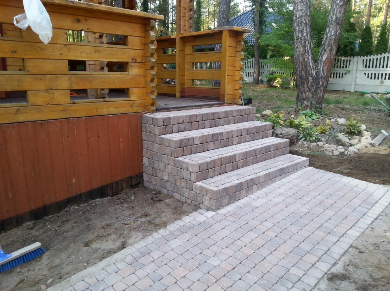 Schody i ścieżka z kostki brukowej prowadzące do drewnianego domku, widoczne świeżo ułożone fragmenty i szczotka do zamiatania.