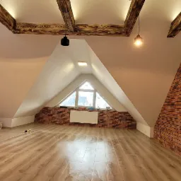 Poddasze po remoncie z oknem trójkątnym, ścianą wyłożoną cegłą klinkierową, drewnianymi belkami stropowymi i lampami wiszącymi, widoczne materiały budowlane i narzędzia.