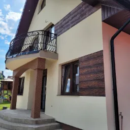 Wykończony dom jednorodzinny z beżową elewacją, dekoracyjnymi panelami imitującymi drewno, żelaznym balkonem i brązowymi elementami wykończeniowymi.