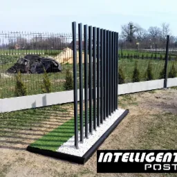 Nowoczesna, minimalistyczna przesłona ogrodowa z pionowych, czarnych profili, osadzona na podstawie z białych kamieni i sztucznej trawy, na tle ogrodzenia z siatki i zieleni