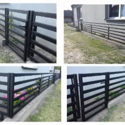 Czarny, poziomy płot metalowy z furtką, zamontowany na betonowej podmurówce, otaczający trawnik i rabatę z kwitnącymi różowymi kwiatami przy szarym domu.