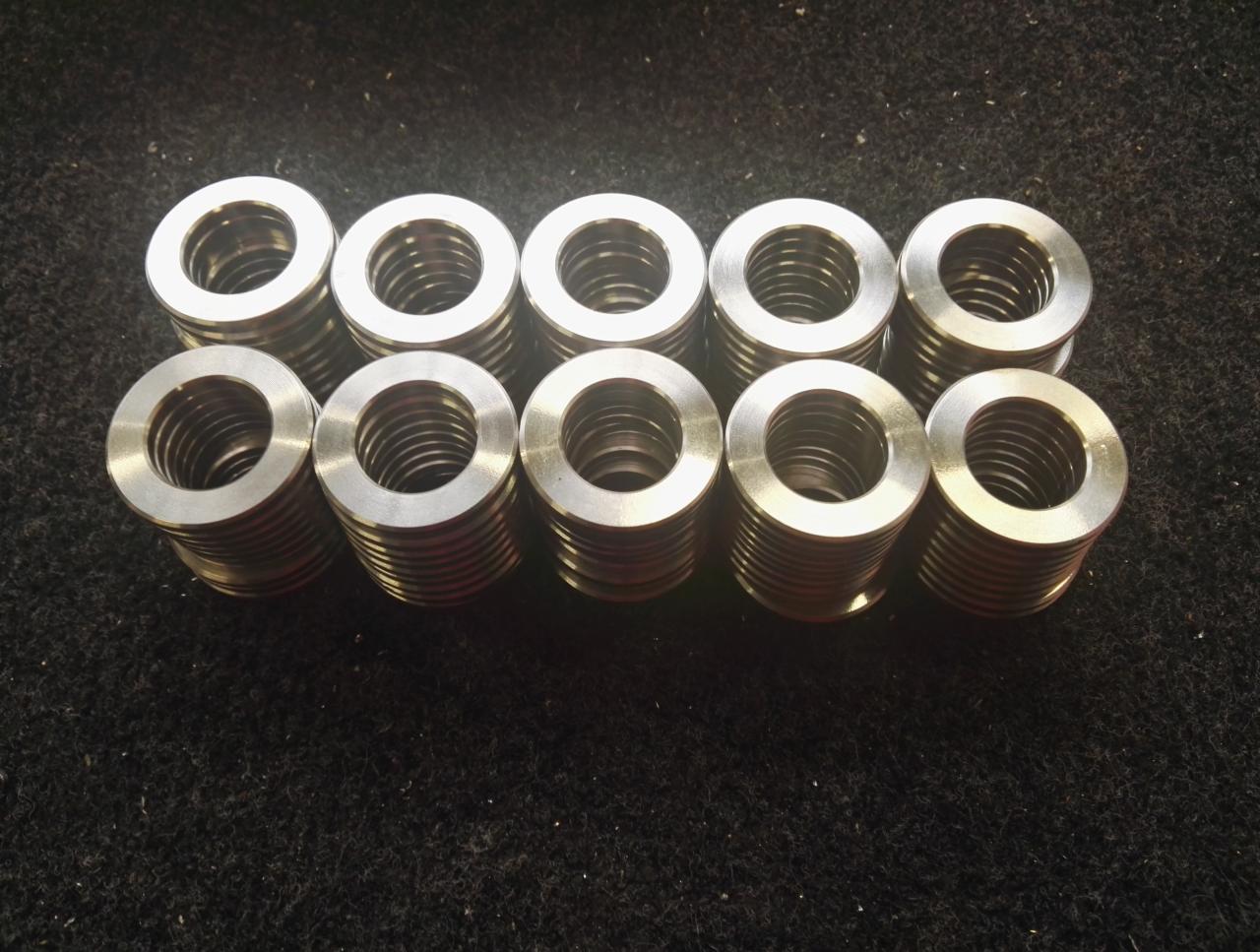Dziesięć identycznych, metalowych tulejek z wewnętrznym gwintem, ułożonych w dwóch rzędach na czarnym, szorstkim podłożu. Widoczne ślady obróbki na powierzchni.