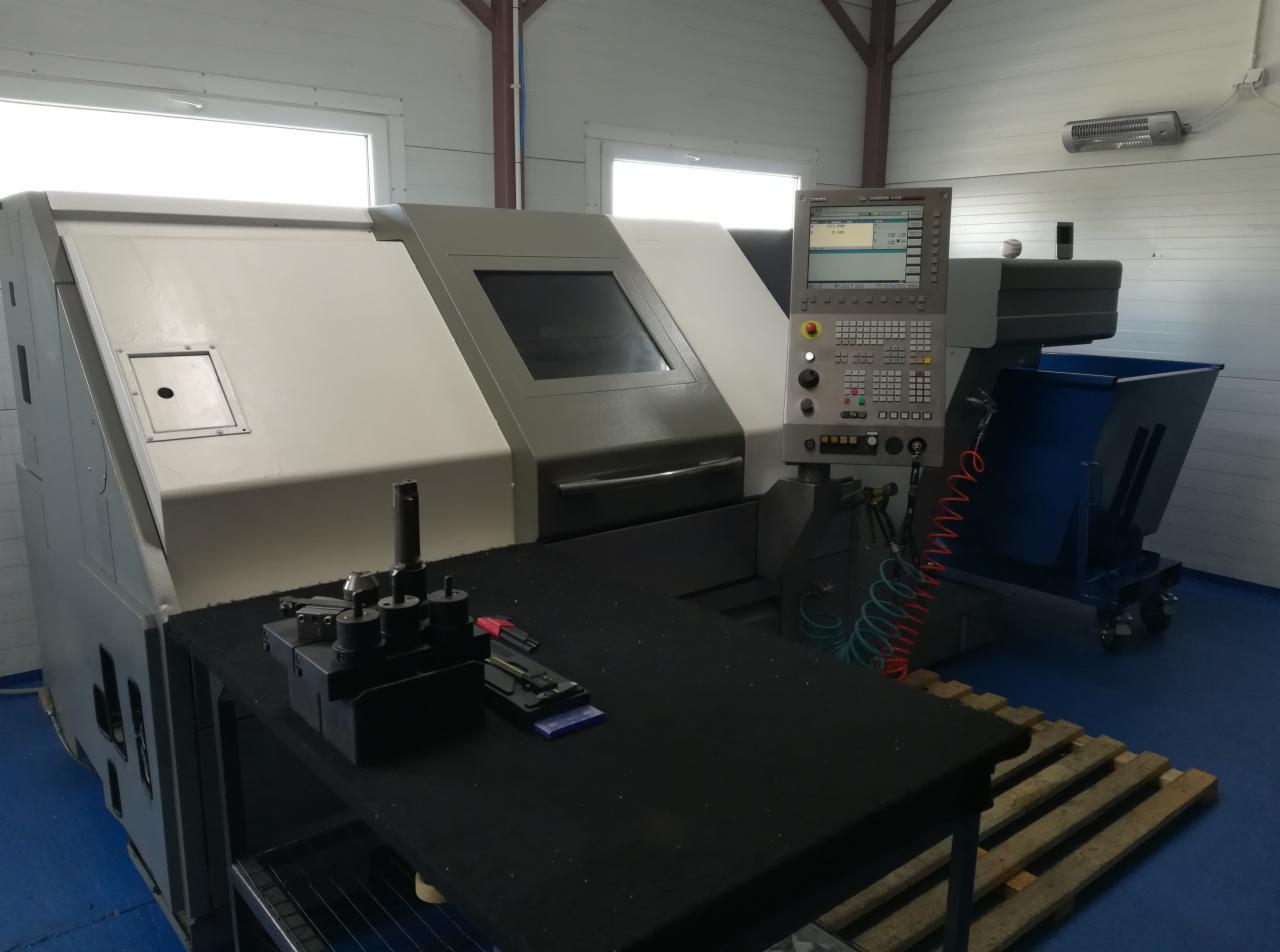 Nowoczesna frezarka CNC z panelem sterowania, narzędziami obok i niebieskim pojemnikiem na odpady na tle hali produkcyjnej.