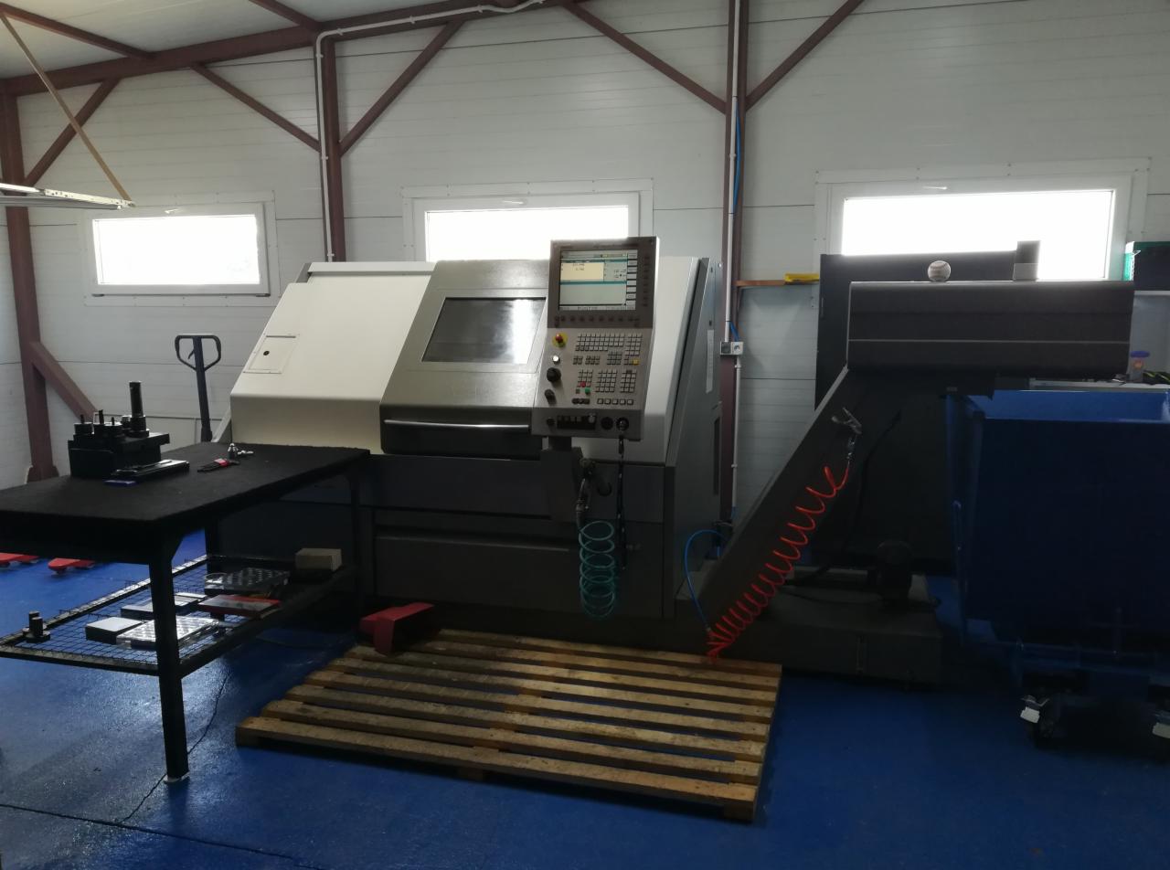 Wnętrze hali produkcyjnej z maszyną sterowaną numerycznie, widoczne okna, paleta pod maszyną, stół z narzędziami i niebieski pojemnik na odpady.