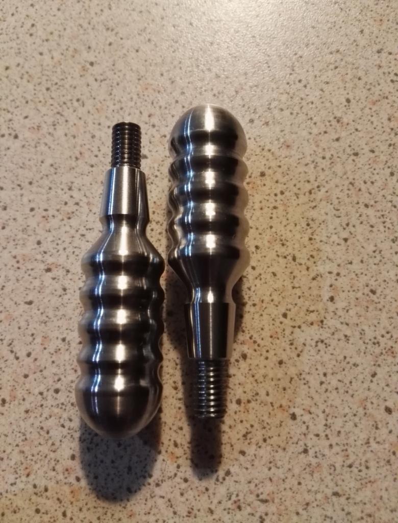 Dwa metalowe uchwyty z gwintem, jeden z kulistą główką i żłobieniami, drugi bardziej smukły, leżące na nakrapianym blacie.