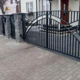 Czarna, metalowa brama wjazdowa z dekoracyjnym łukiem i pionowymi prętami, osadzona między słupkami z szarej kostki, prowadząca do domu z widocznymi schodami wejściowymi.