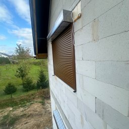 Norbert Wtorkowski - Brązowa roleta zewnętrzna zamontowana na ścianie z białych bloczków, widok z boku, z widocznymi elementami instalacji i fragmentem ogrodu w tle.