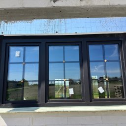 Norbert Wtorkowski - Montaż potrójnego okna w czarnej ramie w surowym murze z betonu komórkowego, widoczne zabezpieczenie folią paroizolacyjną nad oknem oraz naklejki na szybach.