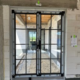 Norbert Wtorkowski - Montaż dwuskrzydłowych okien z czarnymi szprosami w surowym stanie budynku, widoczne bloczki betonowe i elementy konstrukcyjne, zabezpieczenia na szybach.