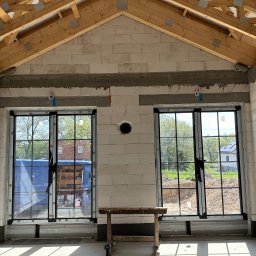 Norbert Wtorkowski - Wnętrze surowego budynku z zamontowanymi oknami z czarnymi szprosami, widoczne drewniane krokwie dachu, ściany z betonu komórkowego z widoczną instalacją elektryczną, na pierwszym planie...