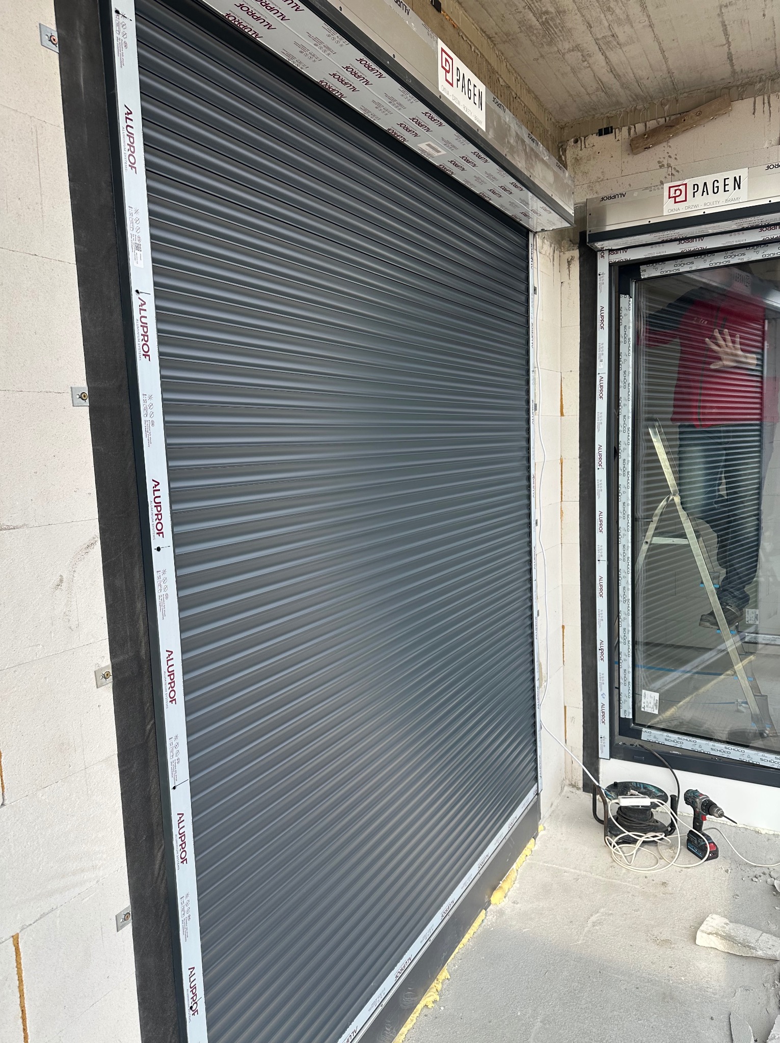 Montaż zewnętrznej rolety aluminiowej w nowym budynku, widoczne materiały izolacyjne, narzędzia i drabina oparta o okno. Roleta w kolorze antracytowym z widocznym logo producenta na ramie.