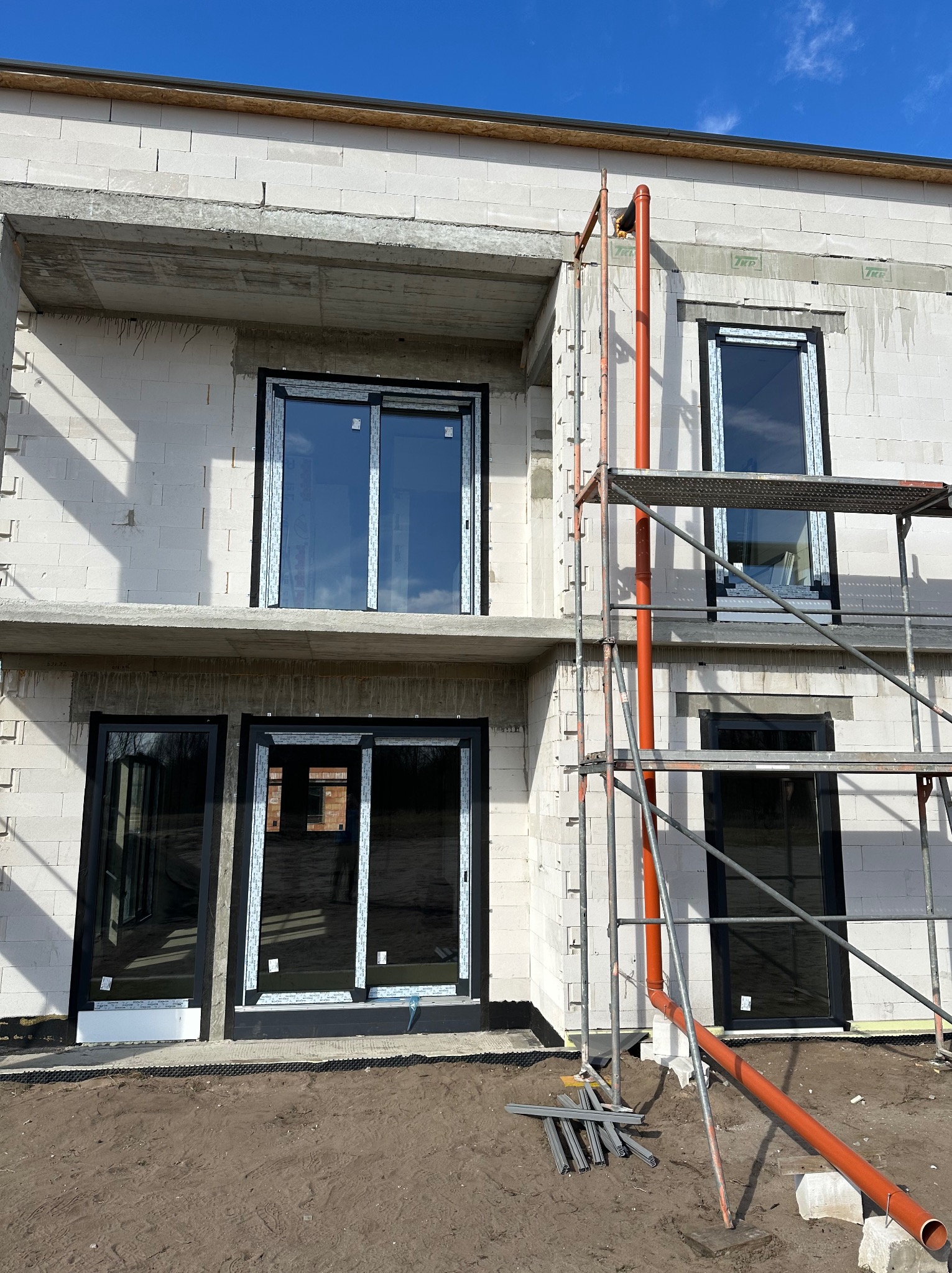 Surowy stan budynku w trakcie montażu okien z czarnymi ramami, widoczne rusztowanie i elementy instalacji odprowadzającej wodę.