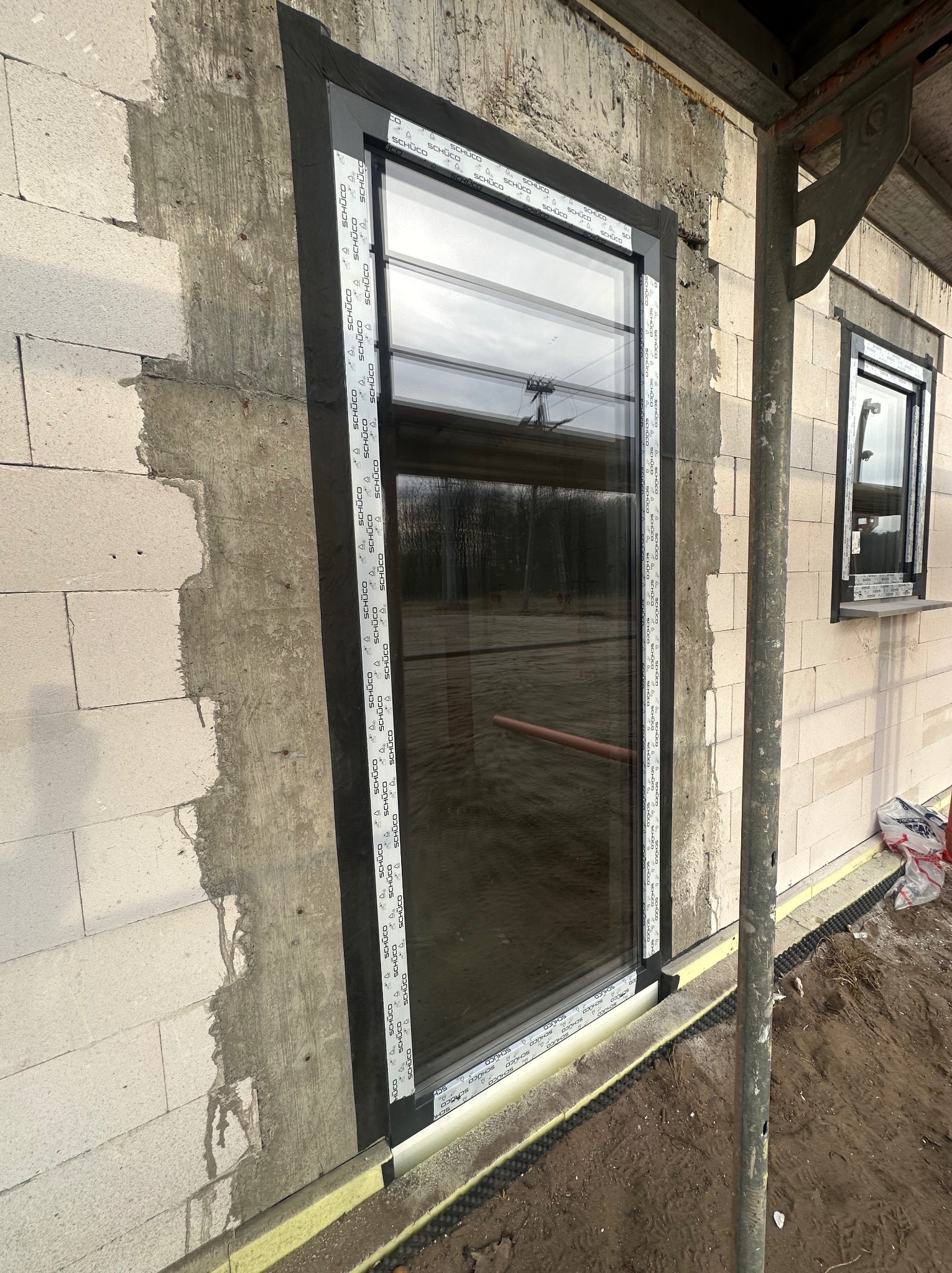 Montaż okna z czarną ramą w surowym budynku z bloczków, widoczne rusztowanie i folia hydroizolacyjna przy fundamencie.