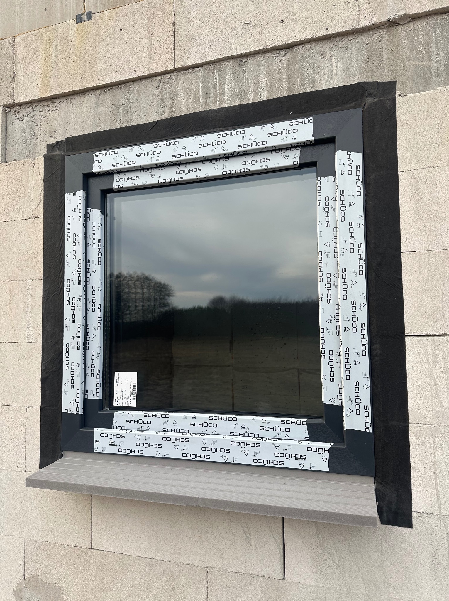 Nowe okno w trakcie montażu w surowym murze z betonu komórkowego. Widoczne taśmy ochronne z logo producenta na ramie i czarna folia uszczelniająca. Na zewnątrz widok na drzewa i pochmurne niebo.