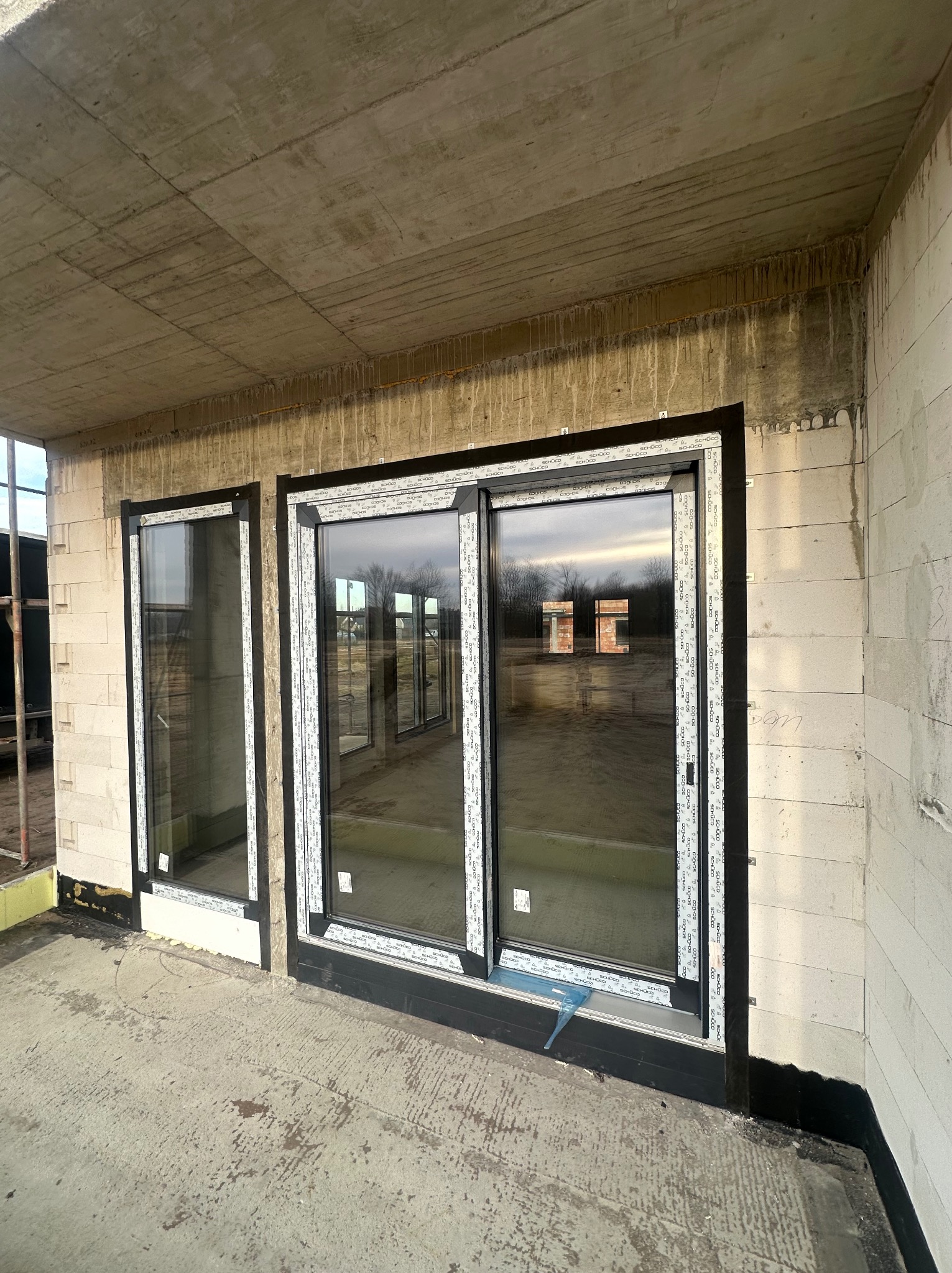 Montaż okien w surowym stanie budynku, widoczne dwa okna z czarnymi ramami i białymi listwami, betonowy strop i ściany z bloczków.