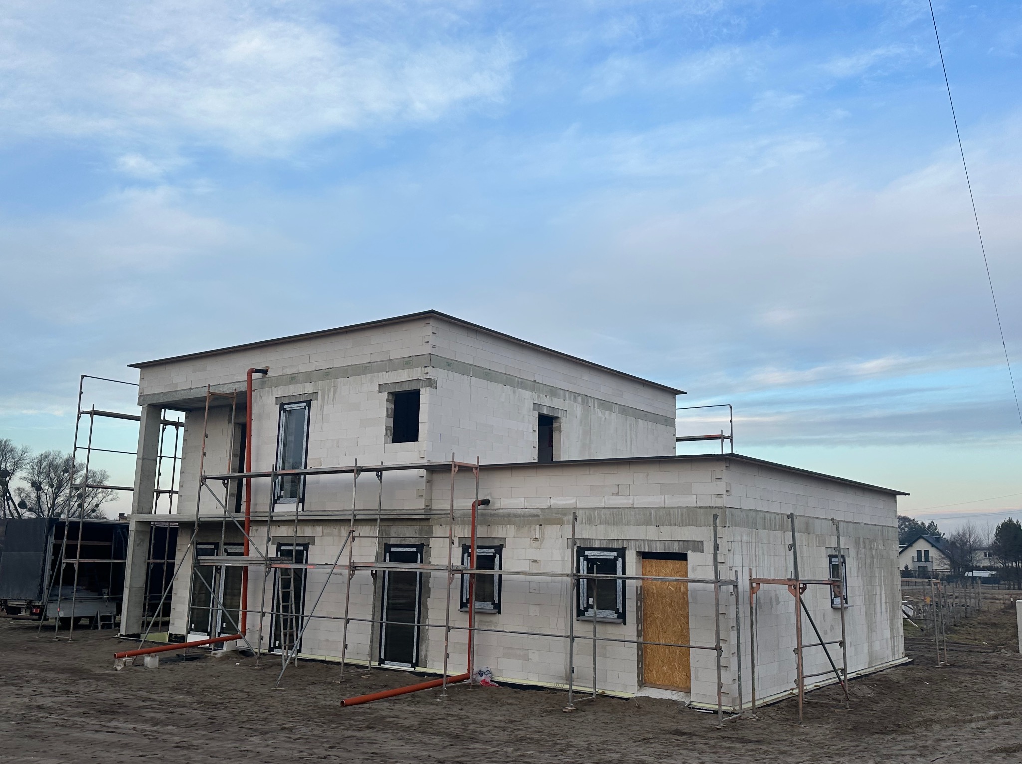 Surowy, dwukondygnacyjny budynek z białych bloczków w trakcie budowy, z widocznymi rusztowaniami, tymczasowo zabezpieczonymi otworami okiennymi i drzwiami, na pierwszym planie rury PCV w kolorze...