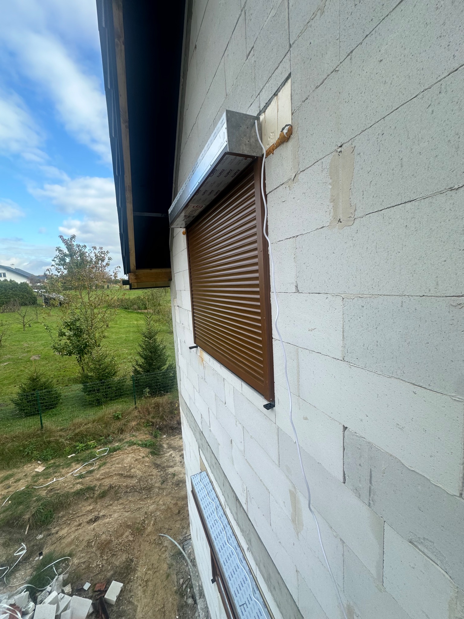 Brązowa roleta zewnętrzna zamontowana na ścianie z białych bloczków, widok z boku, z widocznymi elementami instalacji i fragmentem ogrodu w tle.
