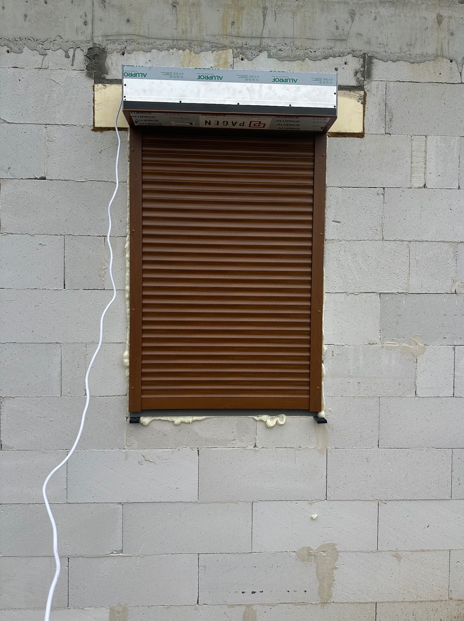 Brązowa roleta zewnętrzna zamontowana w oknie budynku z białych bloczków, widoczna piana montażowa i przewód elektryczny.