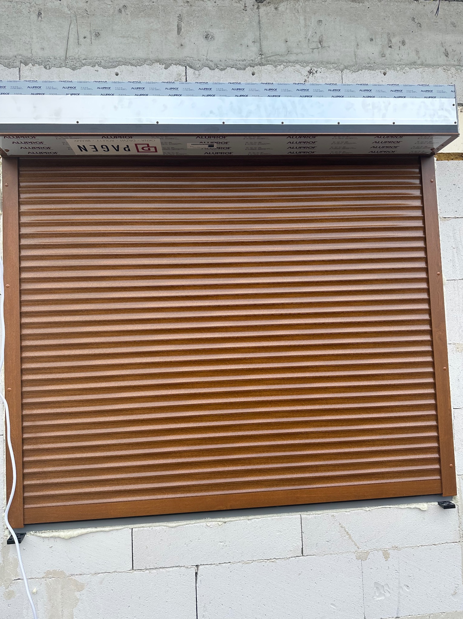 Zamknięta, brązowa roleta zewnętrzna na oknie w trakcie montażu, widoczna na tle surowych, białych bloczków betonowych i z fragmentami pianki montażowej.