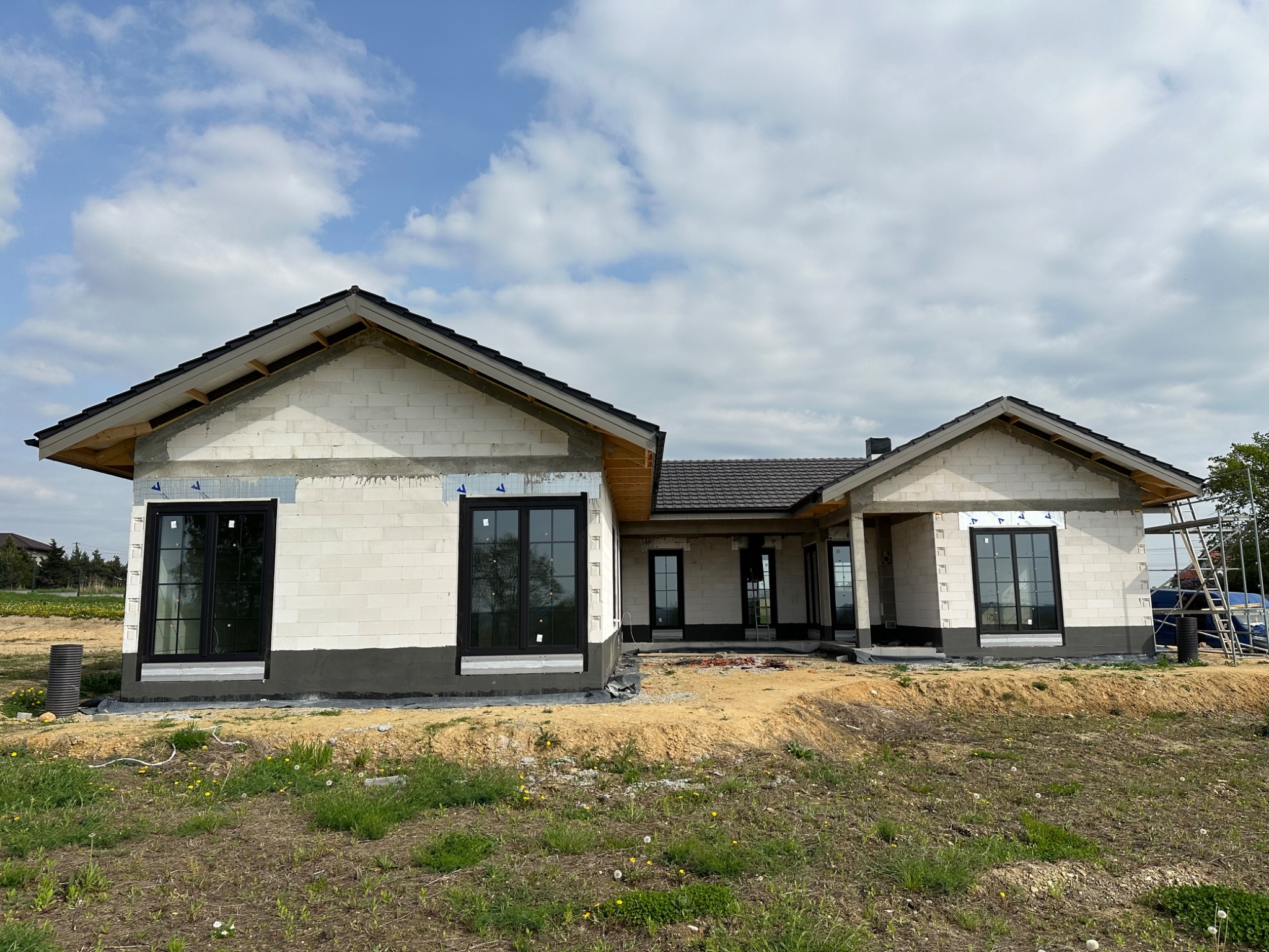 Dom jednorodzinny w trakcie budowy, widoczne czarne okna PCV w surowej elewacji z bloczków, przygotowane do dalszych prac wykończeniowych.