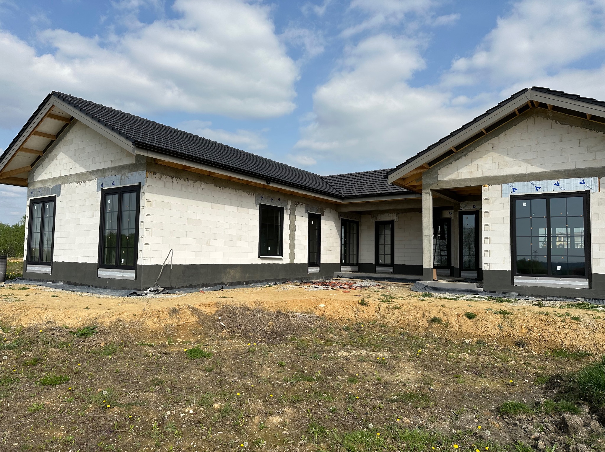 Surowy stan domu jednorodzinnego z czarnymi ramami okiennymi, widoczne bloczki betonowe i czarna dachówka, teren wokół domu jeszcze niezagospodarowany.