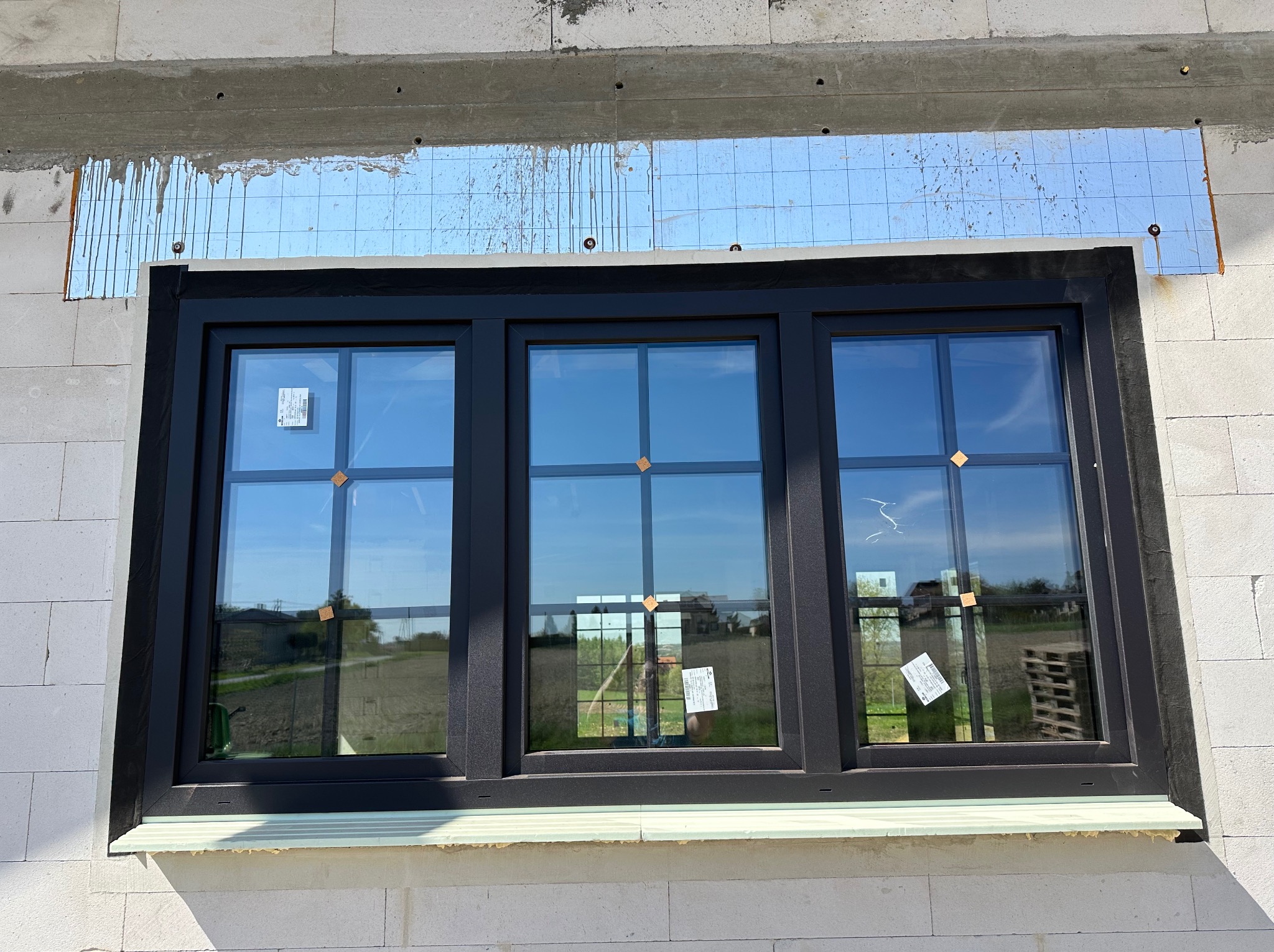 Montaż potrójnego okna w czarnej ramie w surowym murze z betonu komórkowego, widoczne zabezpieczenie folią paroizolacyjną nad oknem oraz naklejki na szybach.