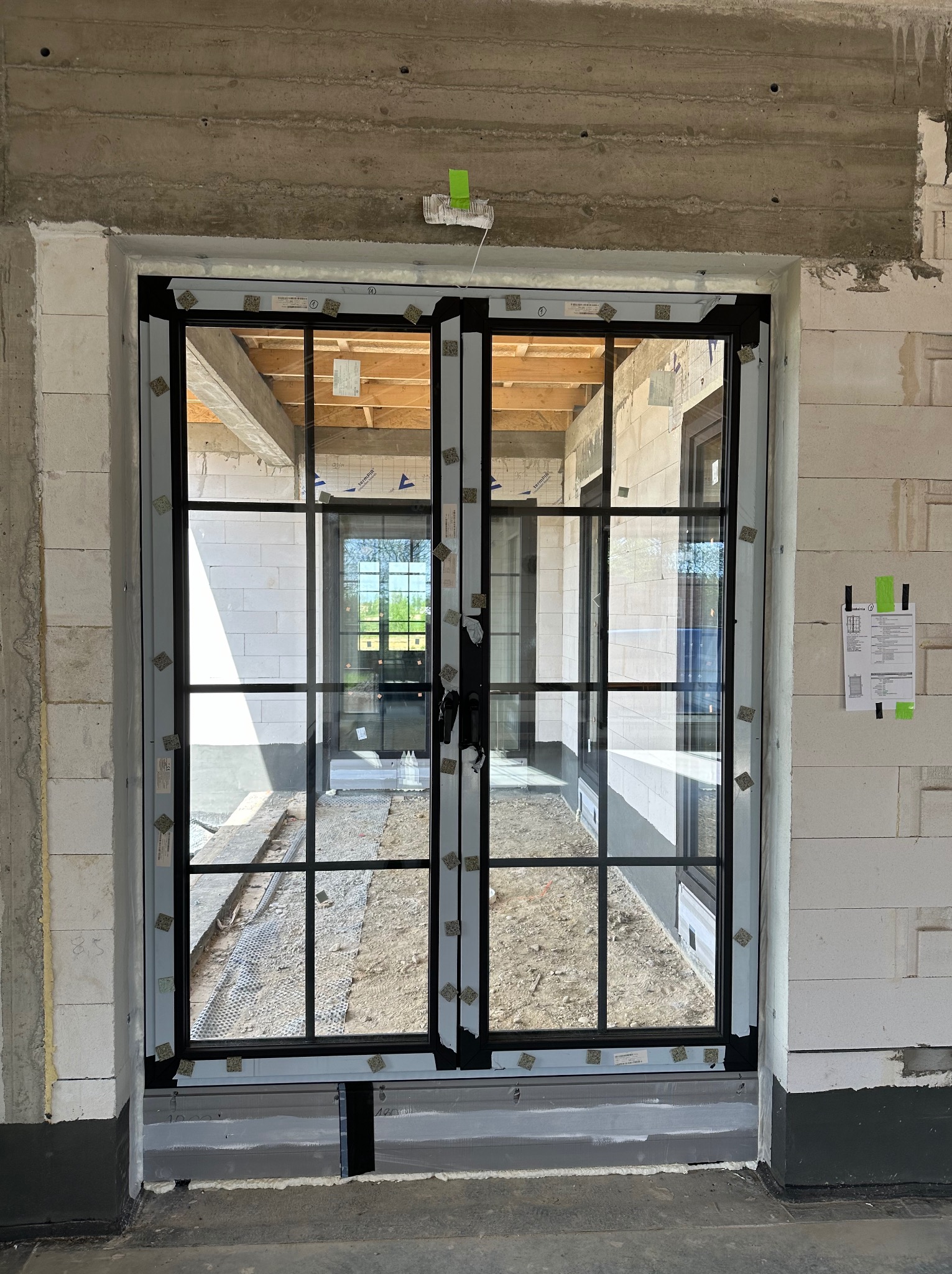 Montaż dwuskrzydłowych okien z czarnymi szprosami w surowym stanie budynku, widoczne bloczki betonowe i elementy konstrukcyjne, zabezpieczenia na szybach.