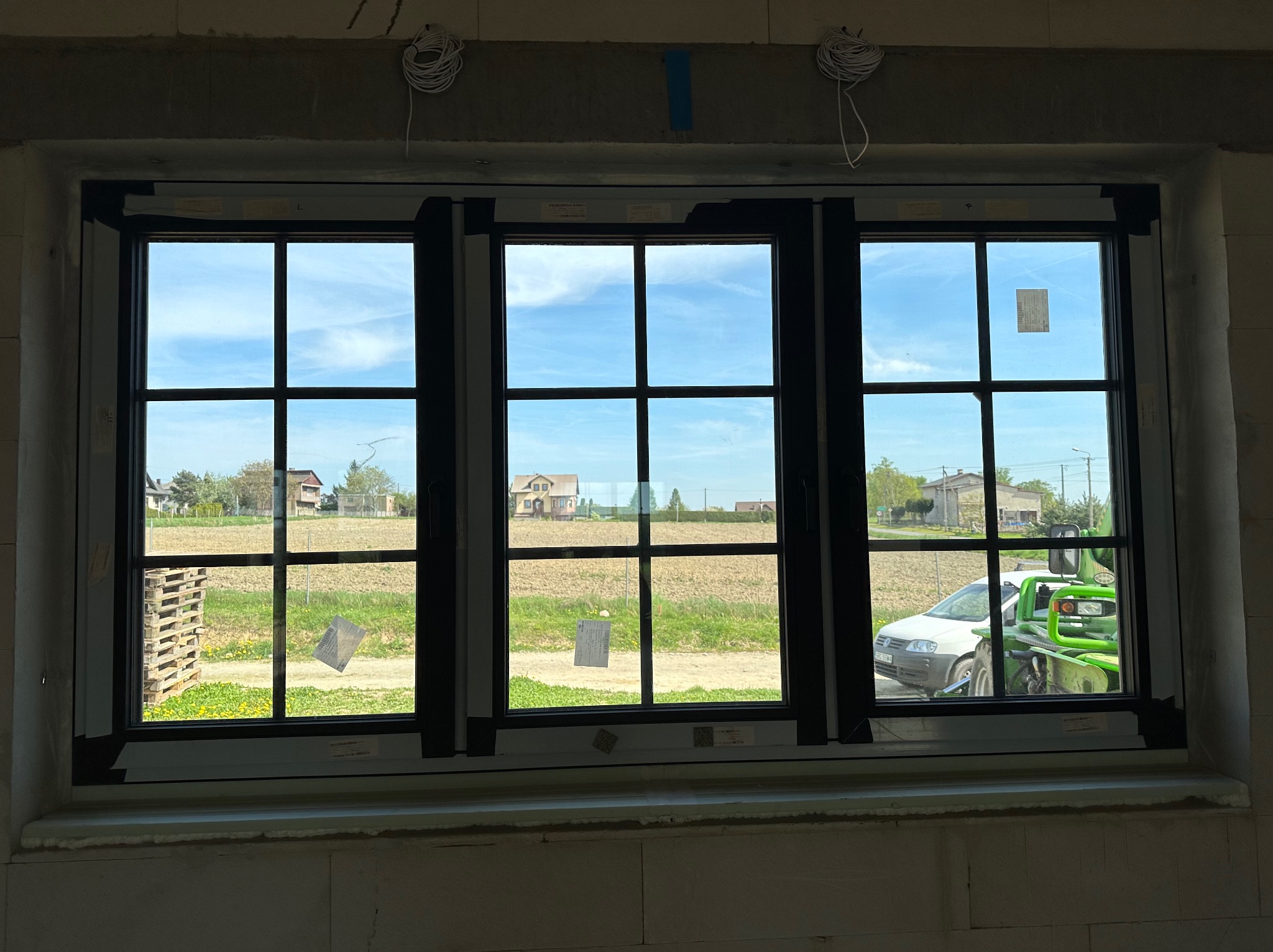 Nowe okno z czarną ramą, zamontowane w surowym murze, widok na pole, domy i częściowo zaparkowany samochód oraz zielony traktor na zewnątrz.