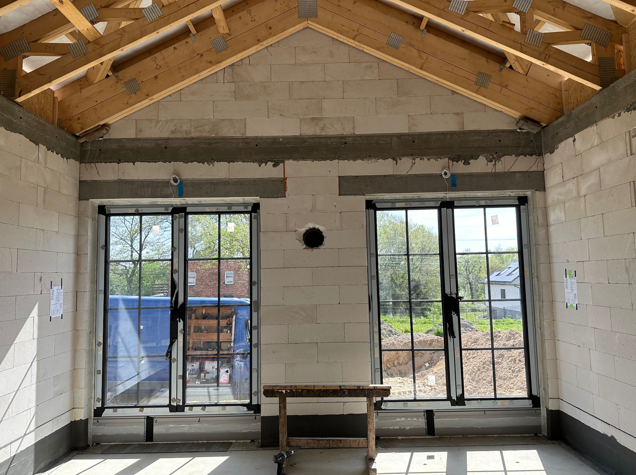 Wnętrze surowego budynku z zamontowanymi oknami z czarnymi szprosami, widoczne drewniane krokwie dachu, ściany z betonu komórkowego z widoczną instalacją elektryczną, na pierwszym planie...