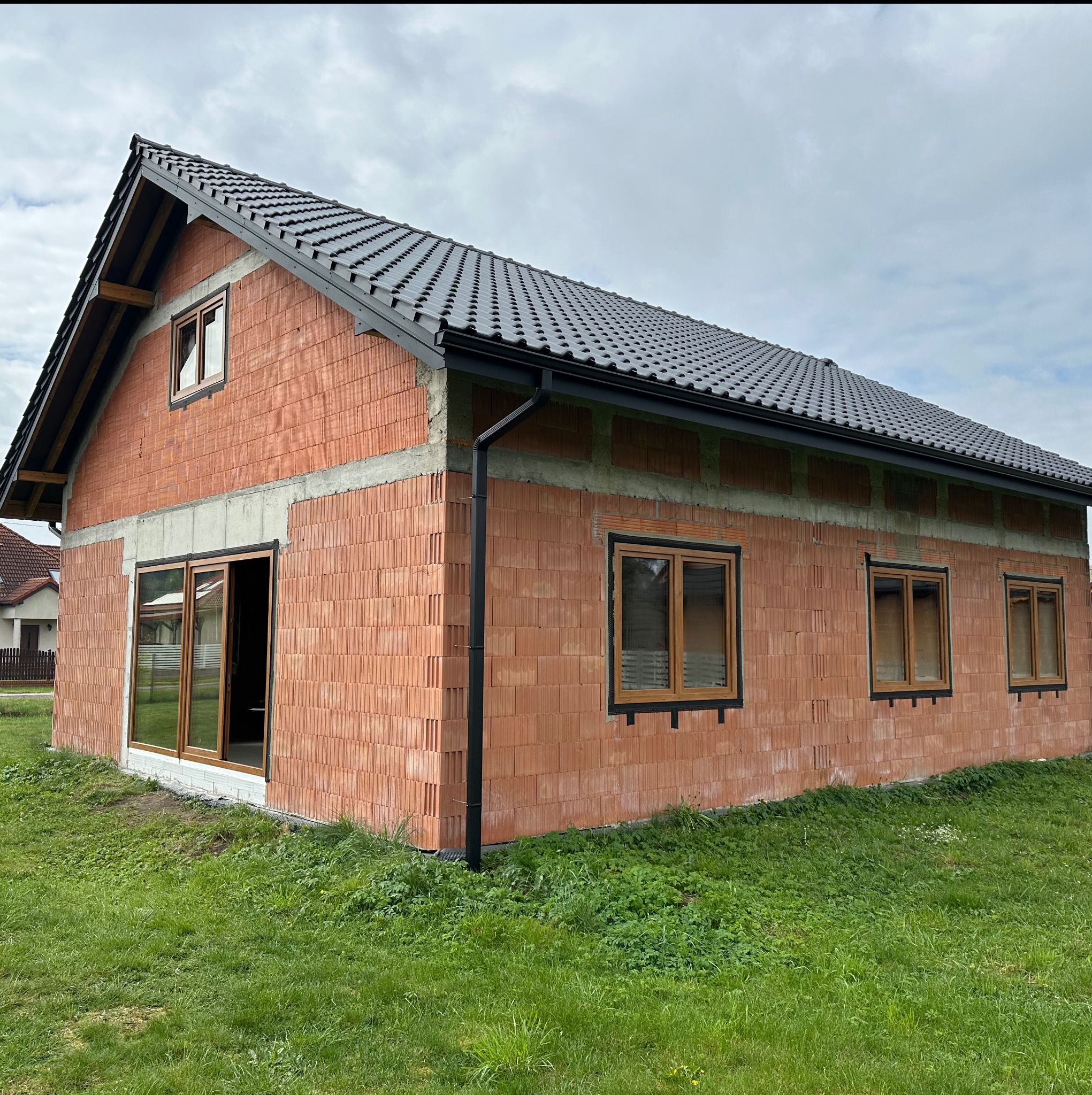 Surowy stan domu jednorodzinnego z czerwonej cegły w Rybniku, z zamontowanymi oknami z brązowymi ramami i czarnymi rynnami, widok z zewnątrz na tle zielonej trawy i pochmurnego nieba.