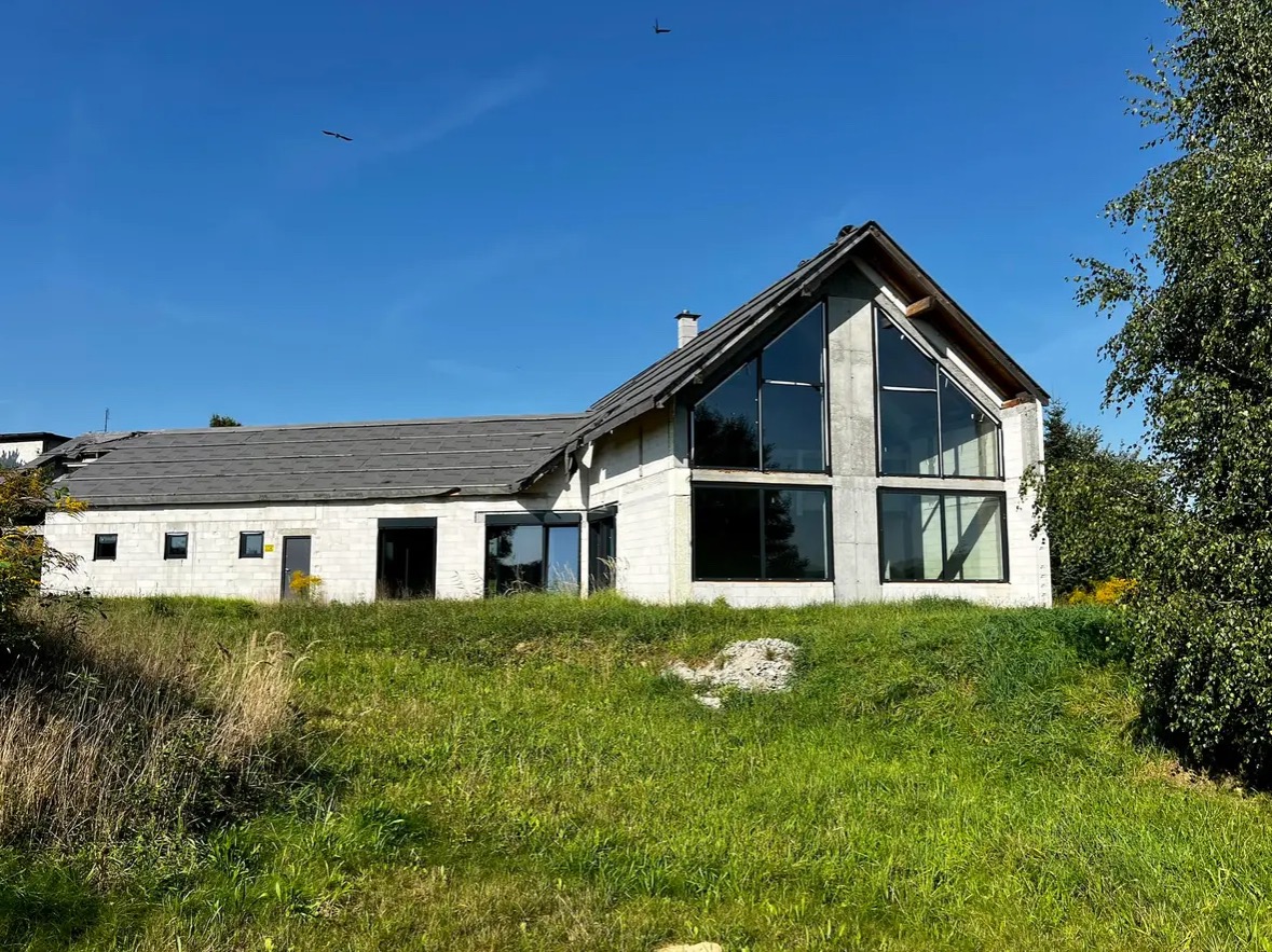 Dom w stanie surowym z zamontowanymi oknami aluminiowymi o dużych przeszkleniach, widok z zewnątrz na tle błękitnego nieba i zielonej trawy.