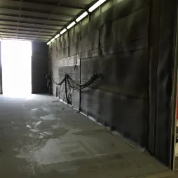 Wnętrze hali z ciemnymi ścianami, prawdopodobnie przygotowanej do piaskowania lub malowania, widoczne przewody i sprzęt BHP przy ścianie.